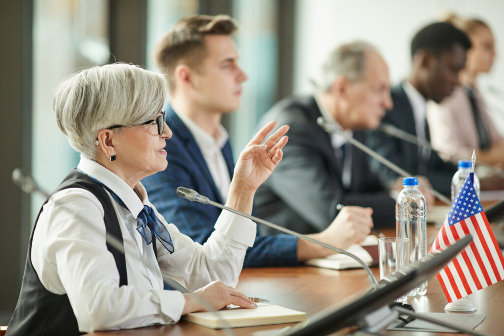 A senior woman with glasses speaks into a microphone at a panel, with other professionals in the background and an American flag on the desk ahead.