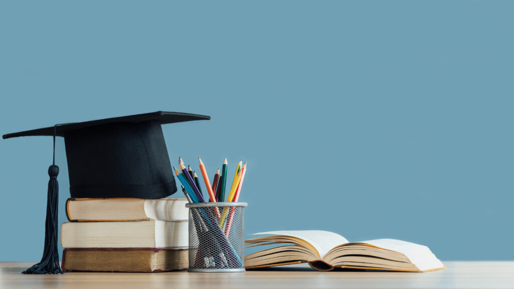 Graduation cap atop a stack of books with a pencil cup and an open book on a desk against a blue background.