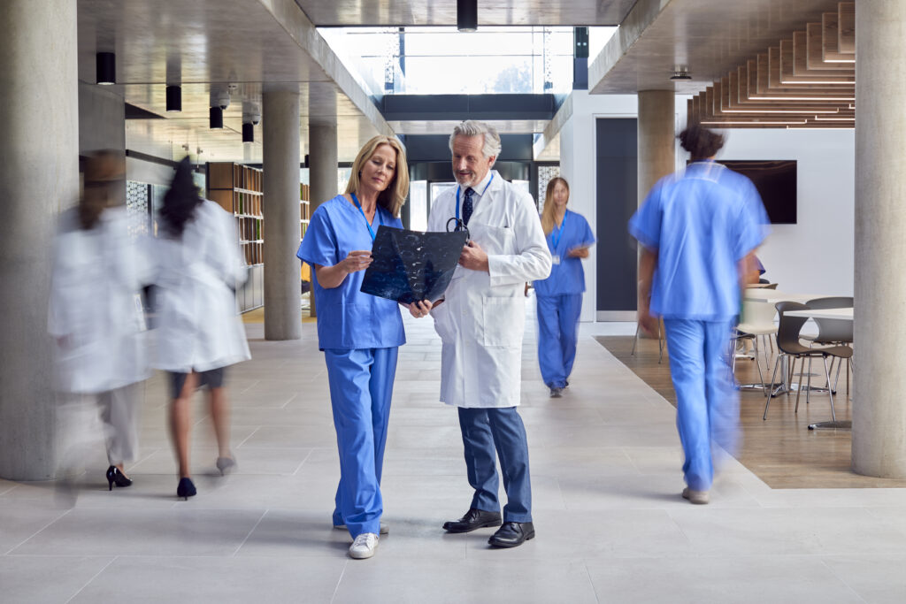 Two medical professionals study a dark X-ray film together in a bright hospital corridor while other staff move by.
