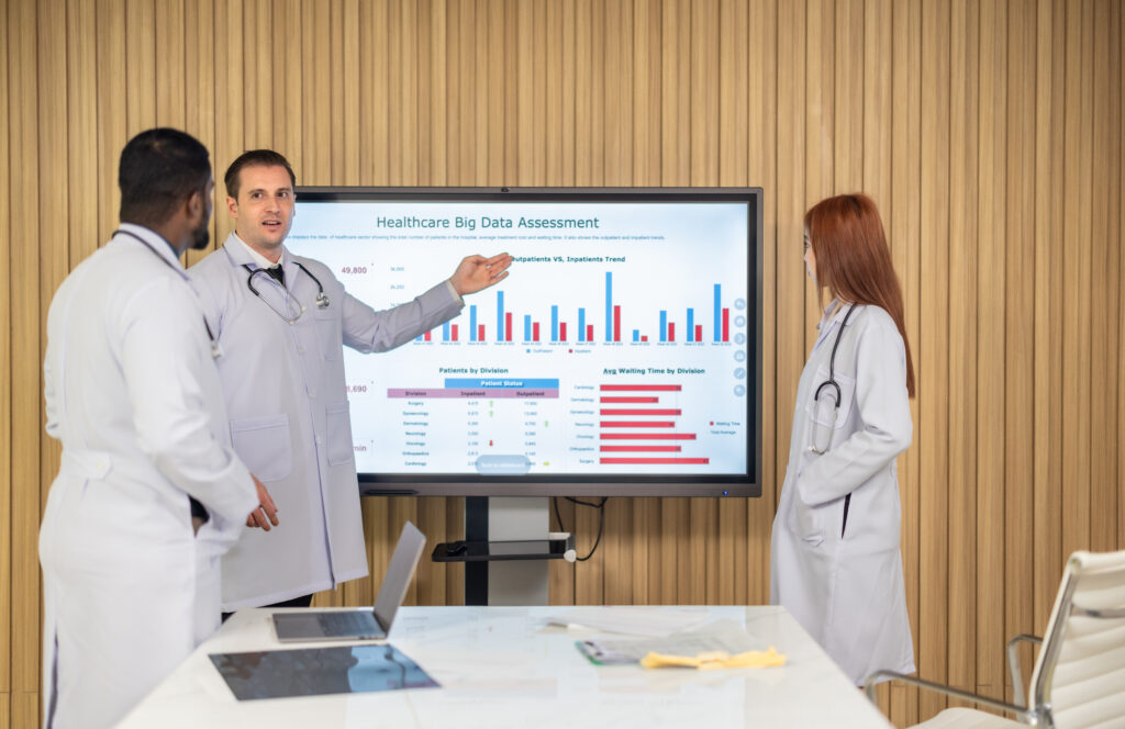 Three doctors in white coats present healthcare data on a large screen with bar charts in a meeting room.