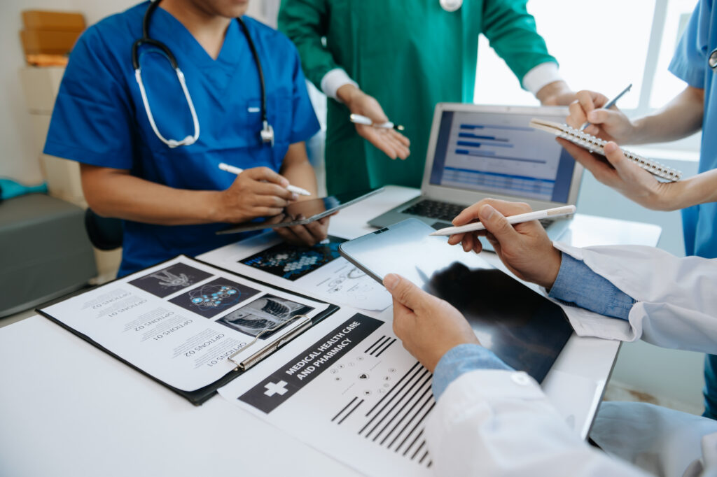 Medical team in blue and green scrubs review charts on a laptop and tablet around a desk.
