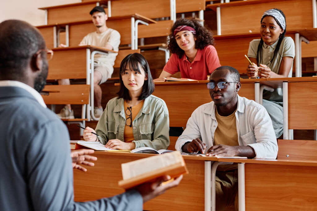 Professor addressing diverse students in a tiered university lecture hall while a student in the foreground takes notes.