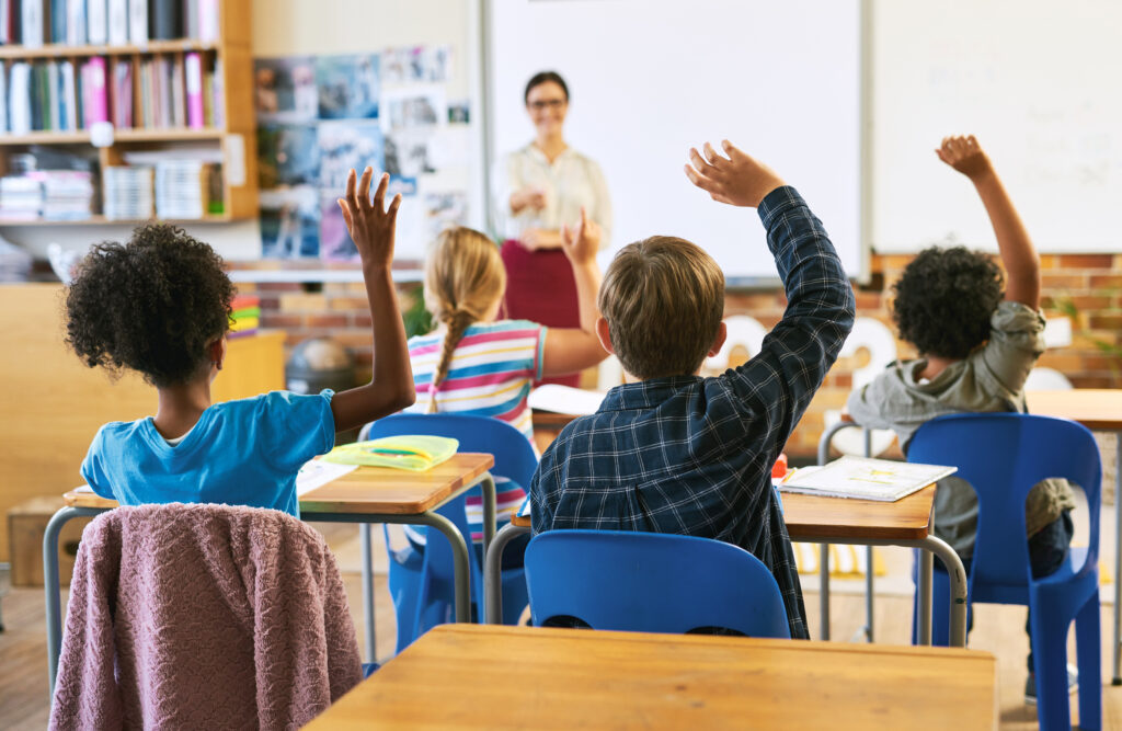Students in a classroom raise their hands as a teacher stands at the front of the room.