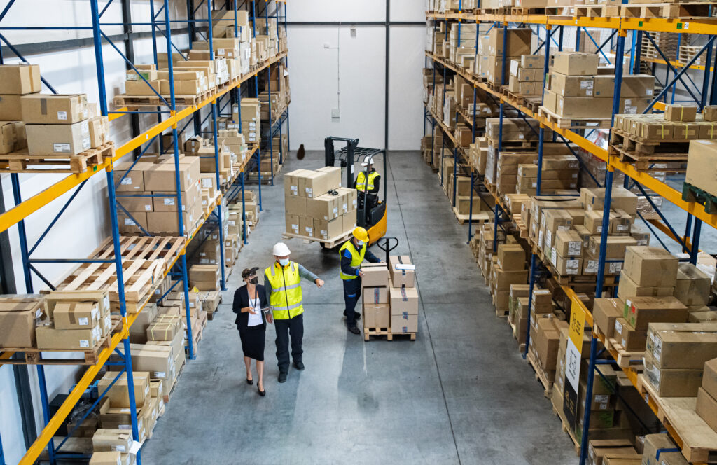 Warehouse aisle with tall blue and yellow racks full of boxes; workers in safety vests handling packages at ground level.