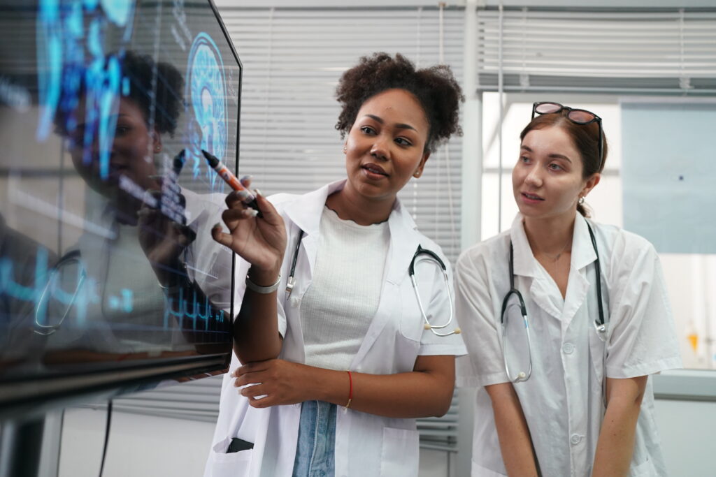 Two female doctors in white coats examine a large touchscreen with blue medical graphics, one writing with a marker while the other observes.